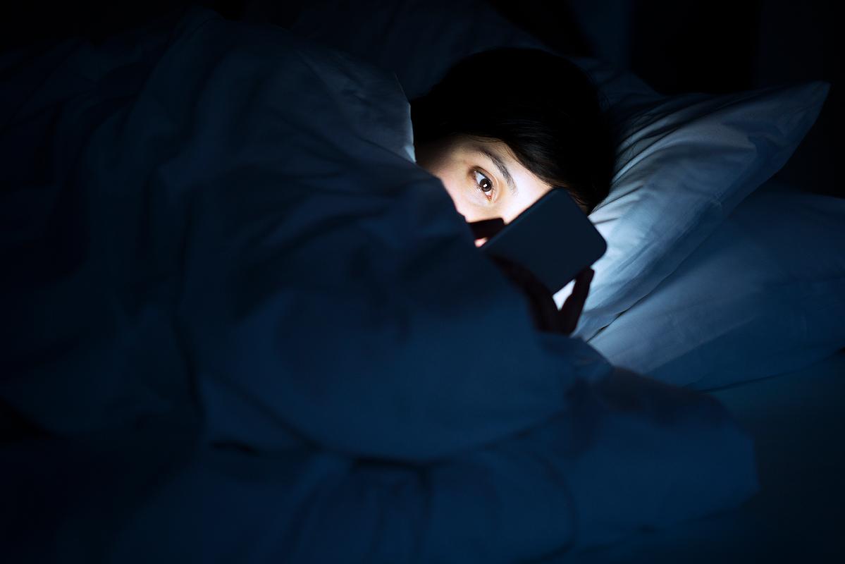 一个女性深夜躺在床上看手机,屏幕白光映出焦虑表情,背景昏暗杂乱