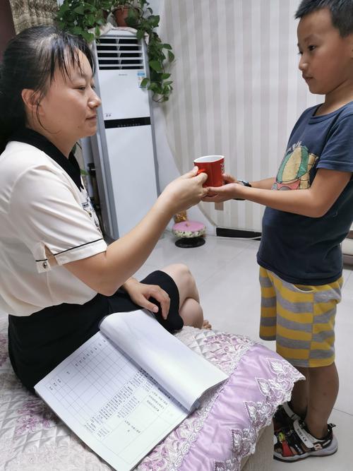 小学低年级男孩站在沙发边给斜靠的妈妈递带吸管的温水杯