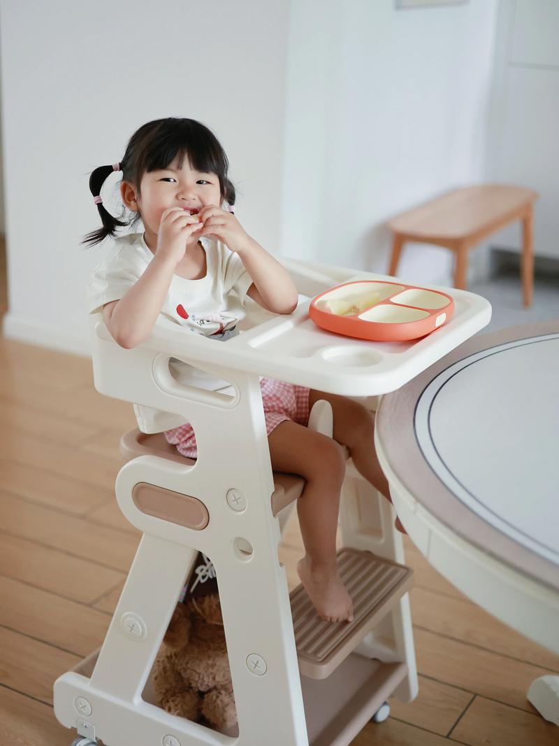 幼儿坐在餐椅里自主进食，面前放着分格餐盘，表情专注